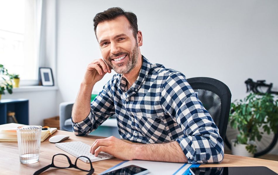 A middle-aged man in a black and white checkered button-up sitting at a desk leaning his head against his hand and smiling