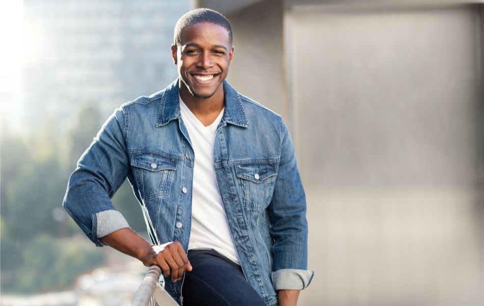 A man in a denim jacket leaning against a rail smiling